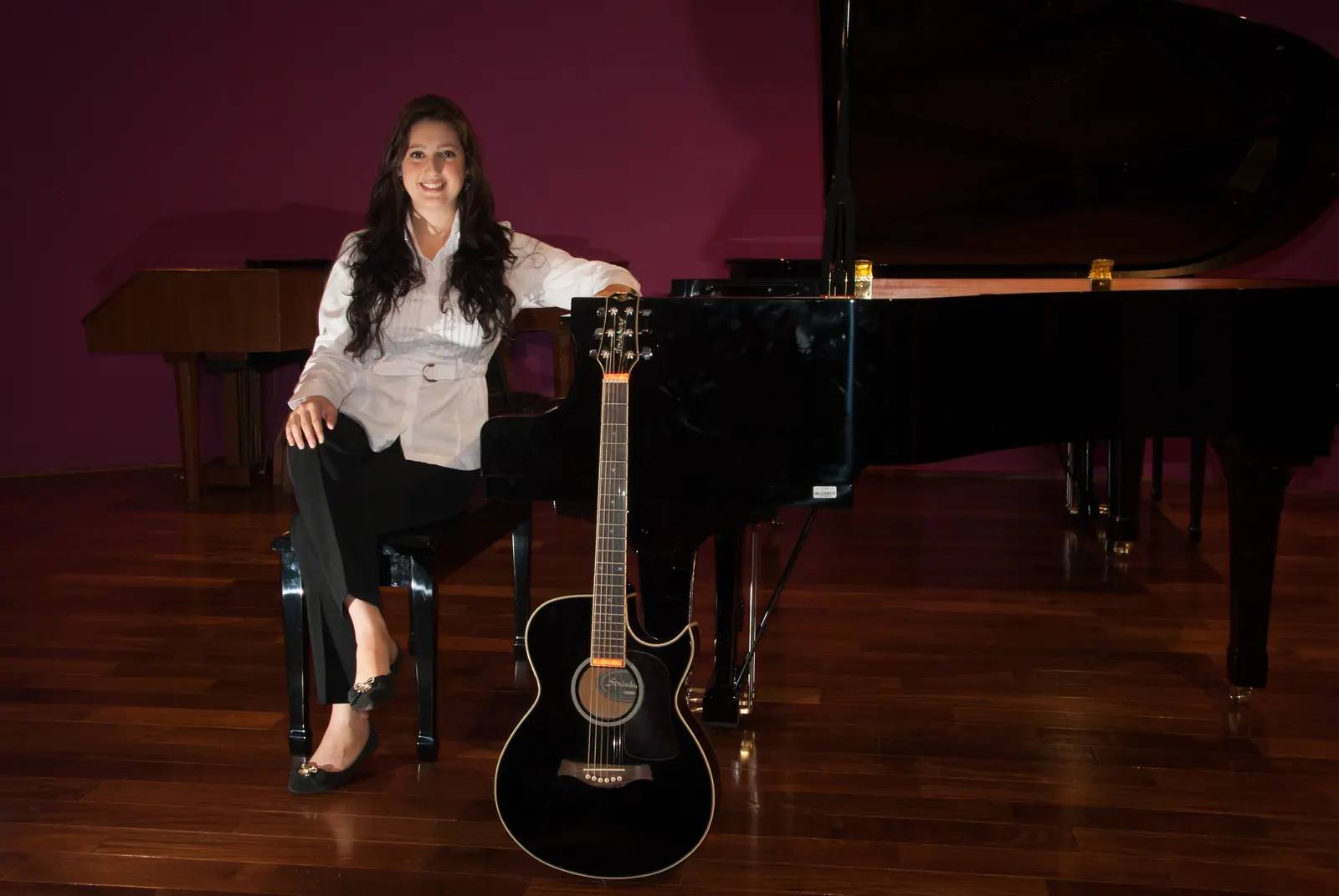 Raquel Gonçalves sentada no banco de um piano de cauda preto, sorrindo, de blusa branca e calça preta, com um violão preto à frente, em uma sala de parede vinho e piso de madeira.