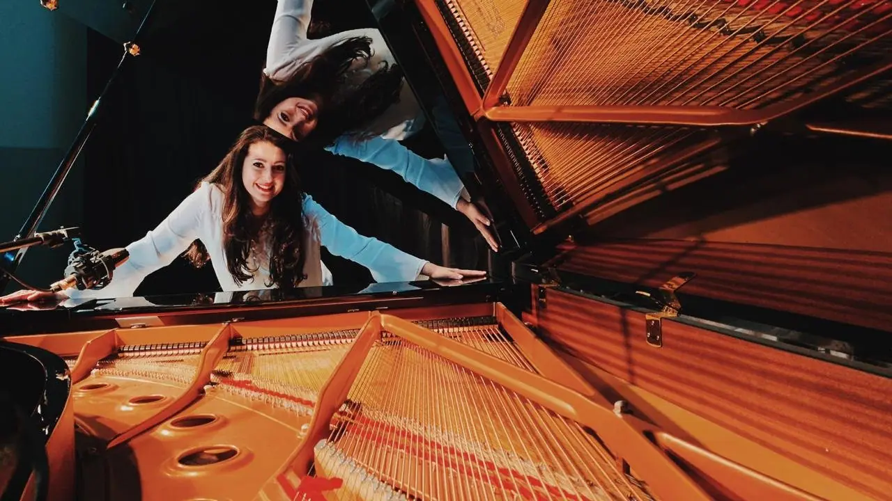 Raquel Gonçalves sorrindo, de blusa branca, com os braços apoiados em um piano de cauda aberto; a tampa do piano reflete a imagem dela de ponta-cabeça.