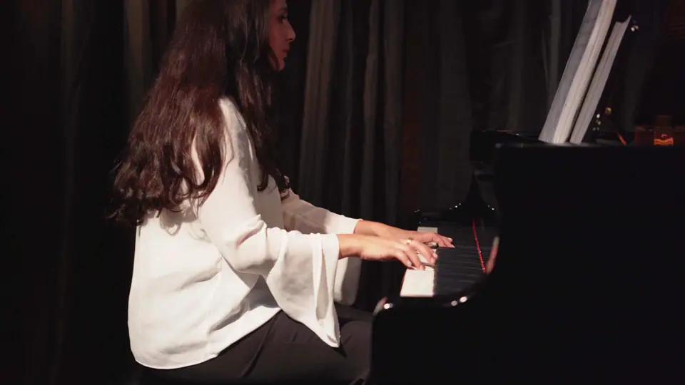 Raquel Gonçalves tocando no palco, de blusa branca, com cortinas escuras ao fundo.