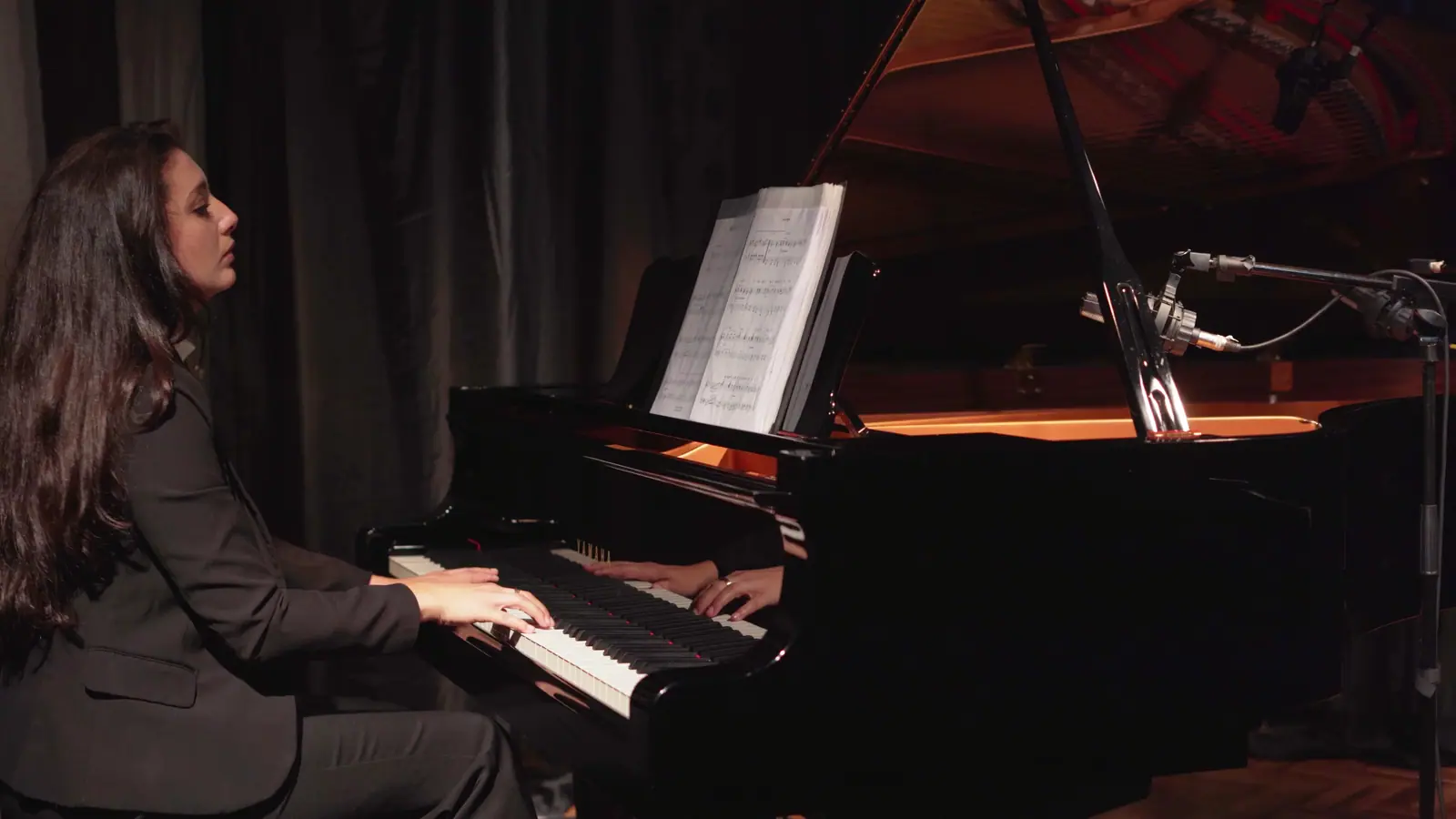 Raquel Gonçalves tocando um piano de cauda no palco, de blazer escuro, com um microfone ao lado do instrumento.