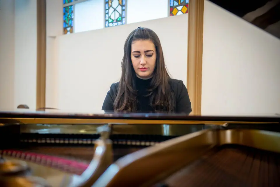 Raquel Gonçalves de olhos baixos, atrás de um piano de cauda, em uma sala com vitrais.