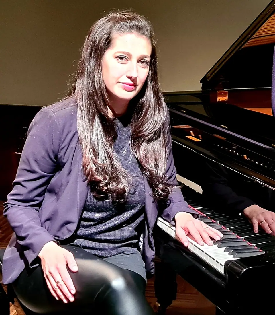 Raquel Gonçalves sentada ao piano, de blazer roxo, com uma das mãos no teclado, olhando para a câmera.