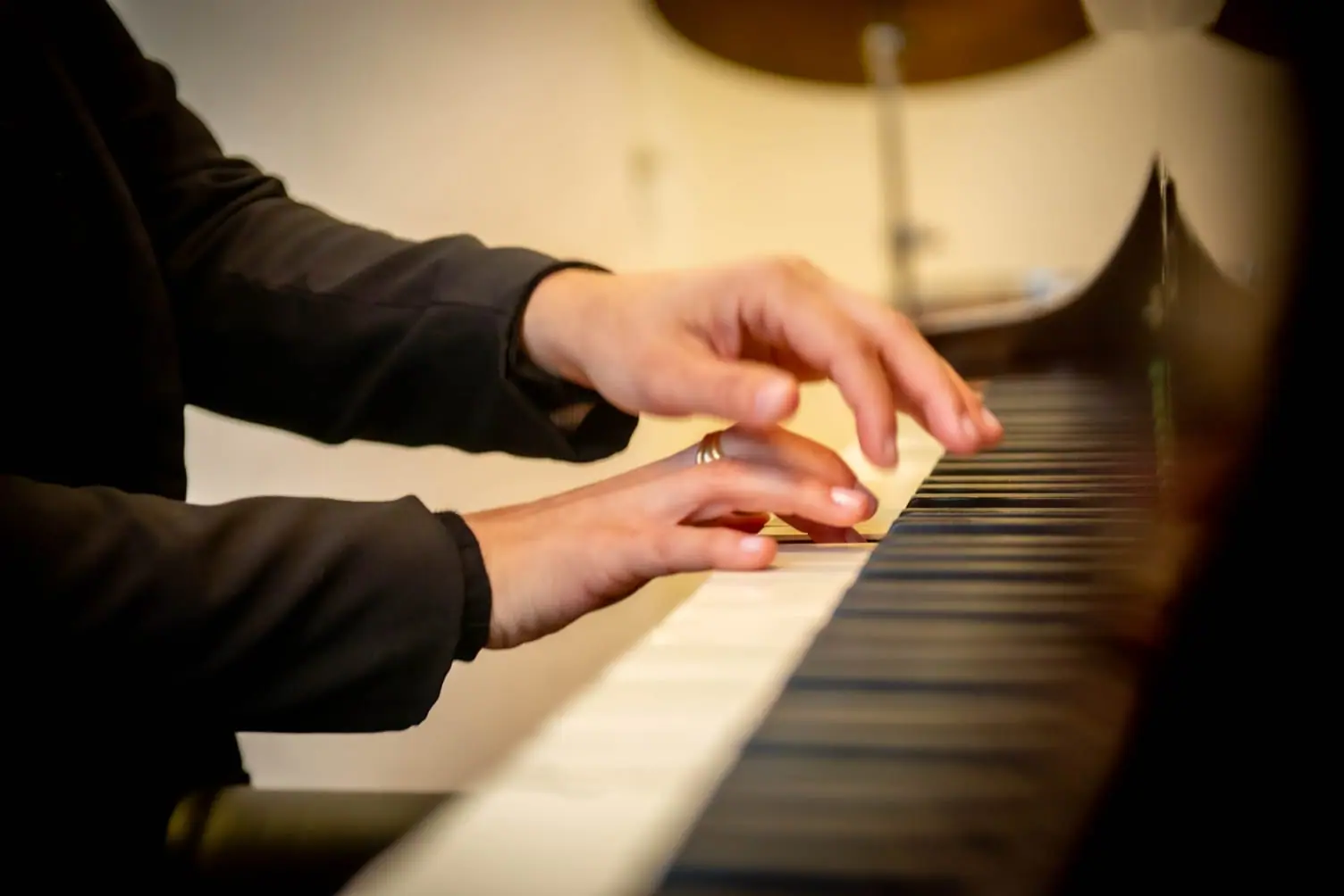 Mãos tocando as teclas de um piano, em close, com a luz quente do ambiente ao fundo.