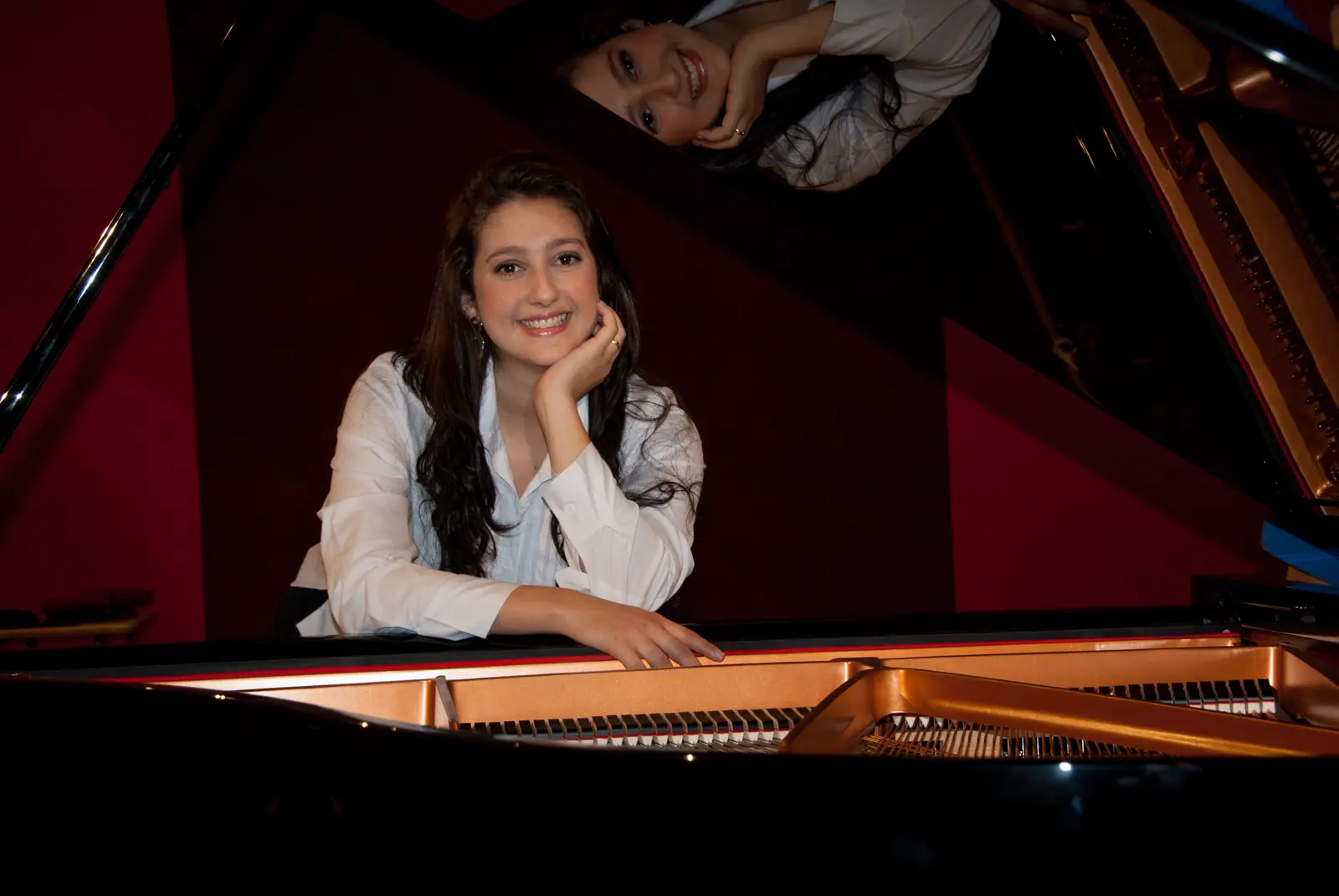 Raquel Gonçalves sorrindo, de blusa branca, com o rosto apoiado na mão, atrás de um piano de cauda aberto; em primeiro plano, as cordas douradas do instrumento, diante de uma parede vermelha.