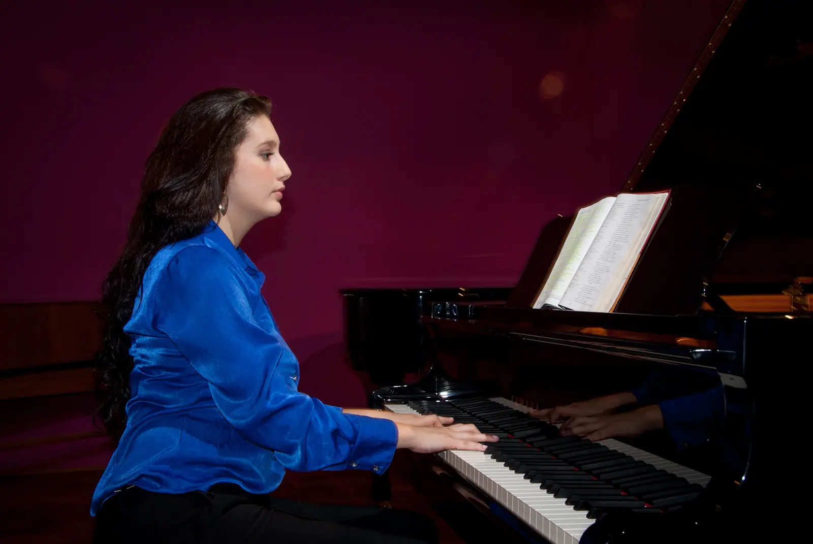 Raquel Gonçalves de blusa azul, tocando piano de perfil, diante de uma parede vinho.